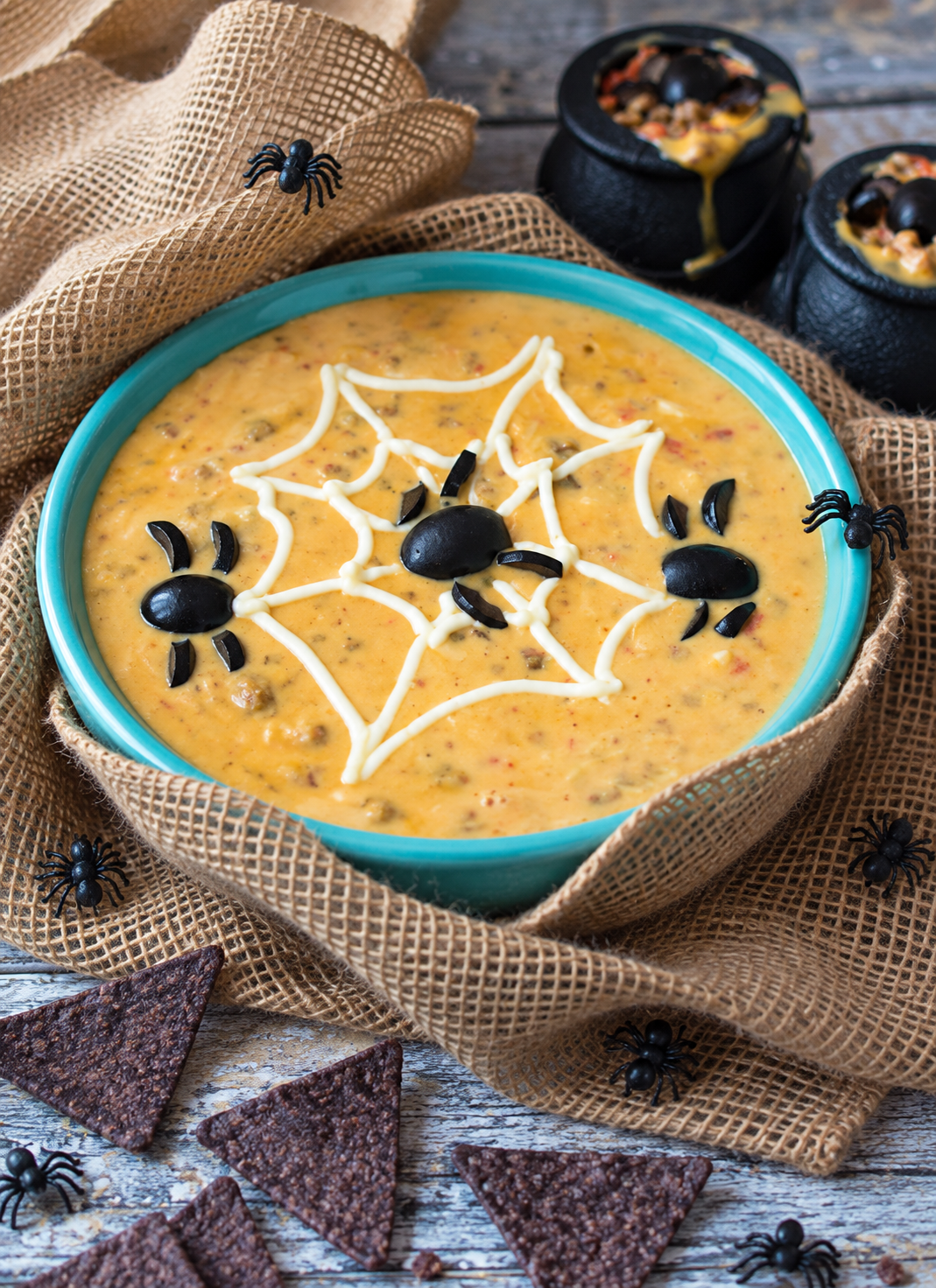 Spooky Spider Sausage Queso in a turquoise bowl decorated with a sour cream spiderweb and black olive spiders.