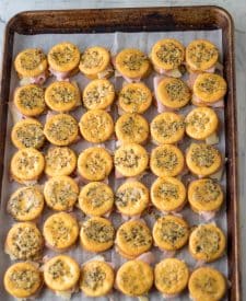 Ritz crackers laid out on a baking sheet with swiss cheese and ham with a cracker on top coated in the butter mixture.