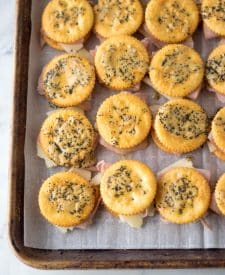 Ritz crackers laid out on a baking sheet with swiss cheese and ham with a cracker on top coated in the butter mixture.