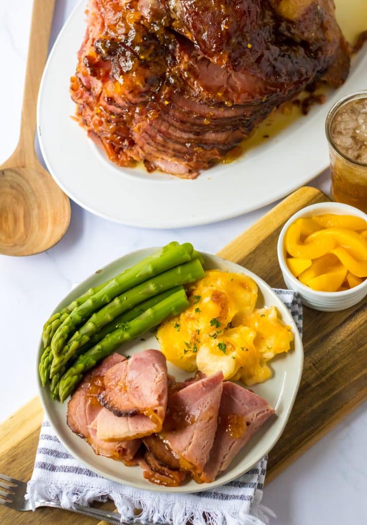Overhead shot of Slow Cooker Hot Honey Peach Glazed Ham on a plate with cheesy potatoes and asparagus