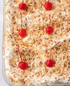 Pina Colada Cheesecake Bars in the pan with whipped topping, toasted coconut and cherries on top