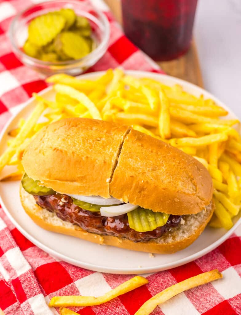 Homemade Copycat McRib sandwich on a plate with golden fries and a red and white napkin underneath.