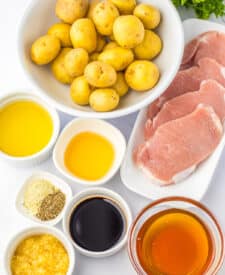 Raw ingredients for honey garlic pork chops laid out on a white surface, including baby potatoes, raw pork chops, garlic, soy sauce, honey, oil, vinegar, and seasoning.