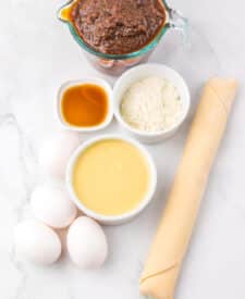 Overhead shot of ingredients for Apple Butter Pie including eggs, flour, vanilla, and apple butter on a white counter.