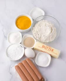 Ingredients for pretzel dogs laid out, including hot dogs, butter, flour, sugar, yeast, and baking soda.