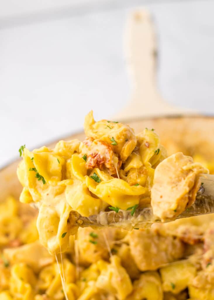 Spoon lifting cheesy chicken tortellini from skillet showing creamy sauce