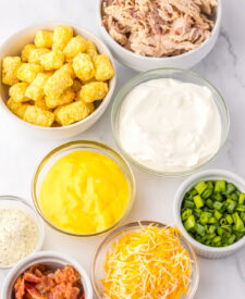 Close-up of ingredients arranged in bowls on a white surface for chicken bacon ranch tater tot casserole.