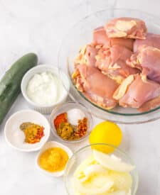Chicken thighs with marinade ingredients and yogurt sauce on white surface
