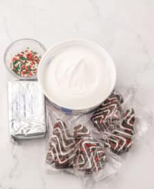 Ingredients for Chocolate Christmas Tree Cake Dip — Little Debbie chocolate Christmas tree cakes, cream cheese, whipped topping, and holiday sprinkles arranged on a festive Christmas table