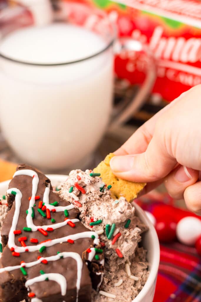 Hand dipping a cookie into creamy Chocolate Christmas Tree Cake Dip topped with red and green sprinkles