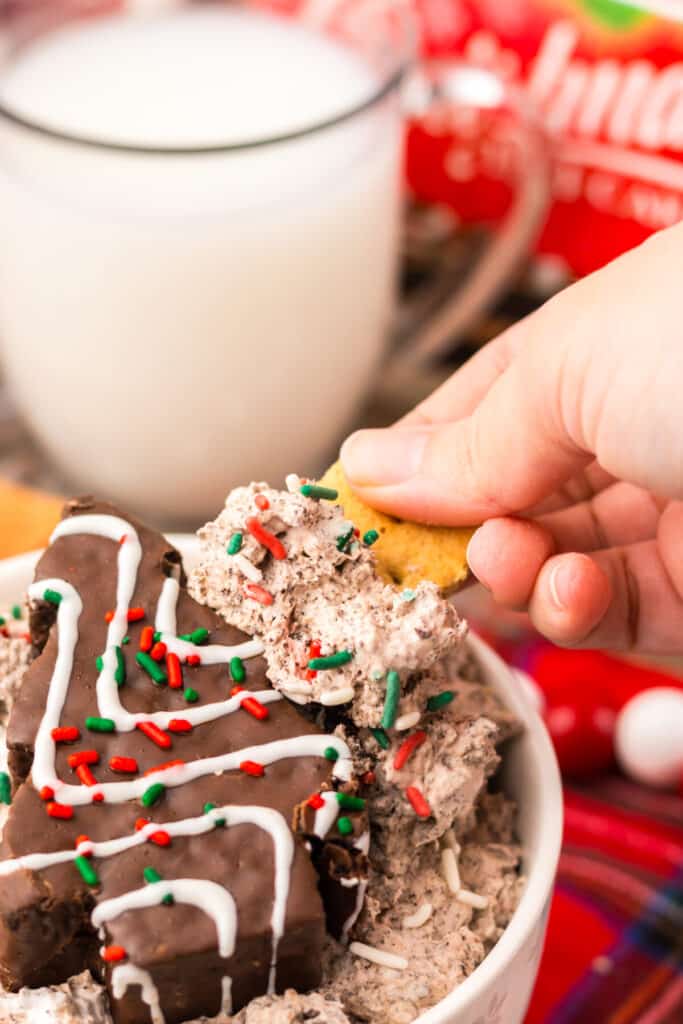 Hand dipping a cookie into creamy Chocolate Christmas Tree Cake Dip topped with red and green sprinkles