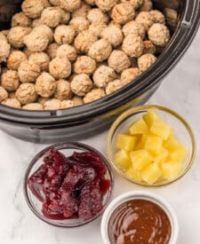 Ingredients for Slow Cooker Cranberry Pineapple Meatballs, frozen meatballs, cranberry sauce, pineapple chunks, BBQ sauce, and fresh parsley laid out on a white counter.