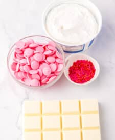 Ingredients for strawberry Cool Whip candy including pink marshmallows, white chocolate, Cool Whip, and sprinkles laid out on a countertop.