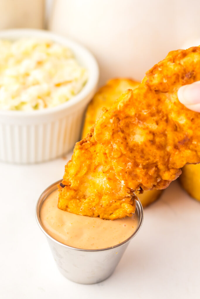 Golden crispy Copycat Raising Cane's chicken tender held above Cane's sauce.